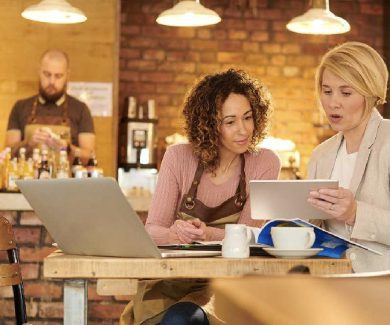 Twee vrouwen in een café-setting tijdens een workshop over financiële grip op je onderneming; een vrouw in een schort (ondernemer) bespreekt samen met een zakelijke adviseur een tablet. Op de achtergrond werkt een barista aan de toonbank